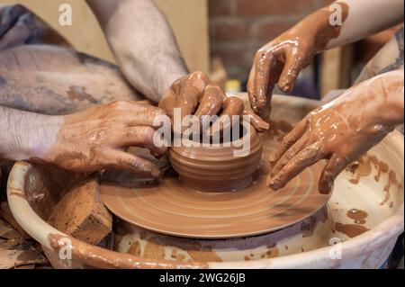 Hände eines Meistertöpfers und eines Kindes, das Geschirr macht. Eine Lektion in einer Töpferwerkstatt. Die Hände eines Töpfchens, der ein Erdenglas auf dem Kreis kreiert. Stockfoto