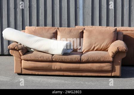 Ein brauner zweiter Hand benutzte die Couch, die draußen auf dem Bürgersteig stand. Ein zusammengerollter Teppich liegt auf dem Sofa. Die Möbel sind alt und abgenutzt. Stockfoto