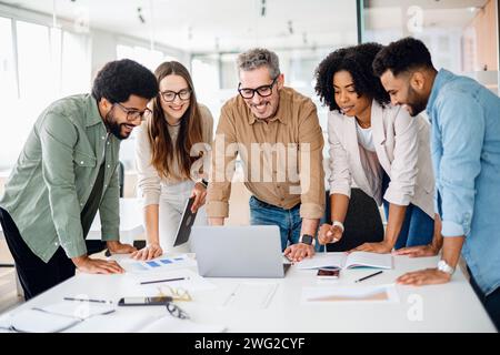 Verschiedene Kollegen treffen sich um einen Laptop mit hellen Ausdrücken, was auf eine erfolgreiche Zusammenarbeit und eine lebhafte Brainstorming-Sitzung in einem gut beleuchteten, modernen Arbeitsbereich und Teamwork-Konzept hinweist Stockfoto