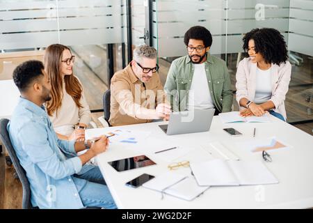 Ein Team führt ein animiertes Gespräch, ein reifer Mann mit Brille gibt leidenschaftlich Gesten, betont einen Punkt, während seine Kollegen aufmerksam zuhören und eine interaktive Brainstorming-Sitzung veranschaulichen Stockfoto