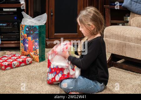 Ein 5-6-jähriges weißes Mädchen sitzt auf dem Boden und reißt am Weihnachtstag Geschenkverpackung von einem Weihnachtsgeschenk ab. USA. Stockfoto