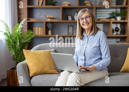 Porträt einer älteren grauhaarigen Frau zu Hause. Eine Frau sitzt auf einem Sofa im Wohnzimmer, lächelt und schaut in die Kamera und arbeitet mit einem Laptop aus der Ferne. Stockfoto