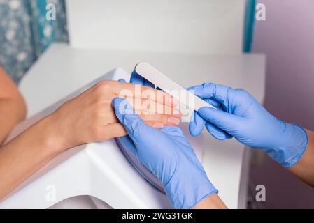 Maniküre. Ein Meister in blauen Handschuhen poliert die Nägel eines Kunden mit einer Nagelfeile in einem Schönheitssalon. Nahaufnahme. Stockfoto