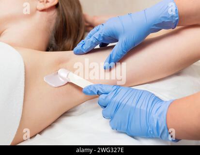 Achselwachsverfahren in einem Schönheitssalon. Meister in Handschuhen trägt Wachs mit einem Holzspachtel auf die Achselhöhle einer Frau auf, um Haare zu entfernen. Stockfoto