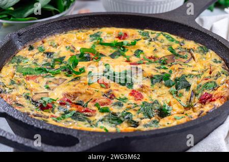 Eierfrittata mit Spinat, gebratenen roten Paprika, Pilzen, Käse und Kräutern, in gusseiserner Pfanne, horizontal, Nahaufnahme Stockfoto