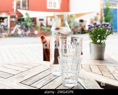 Drei Gläser und eine Karaffe mit Wasser und Eiswürfeln an einem Restauranttisch im Freien. Geringe Schärfentiefe mit fokussierter Brille und Karaffe. Blumen A Stockfoto