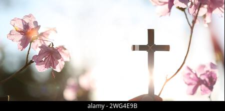 Hintergrund mit starkem Sonnenlicht, das an einem Frühlingstag durch Azaleen-Blüten leuchtet, und einem Christen, der das heilige Kreuz Jesu Christi in seinen Händen hält Stockfoto