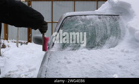 Mit einer Bürste von Schnee befreit eine Hand das Dach und die Windschutzscheibe eines grauen Autos von einer dicken Schneeschicht, die Folgen eines Schnees Stockfoto