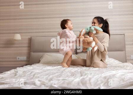 Glückliche Mutter und ihre kleine Tochter spielen mit Hasenspielzeug im Schlafzimmer, Familie hat Spaß Stockfoto