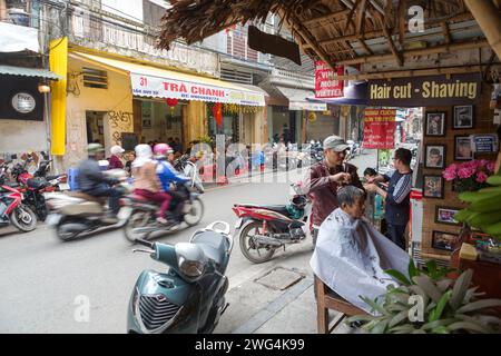 Hanoi, Vietnam, Friseurladen mit Friseur, der Haare eines Kunden auf dem Gehweg schneidet. Stockfoto