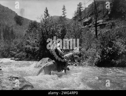 BullChief beim ford, 1905. BullChief trägt eine Kriegshaube und überquert seichte Stromschnellen zu Pferd. Stockfoto