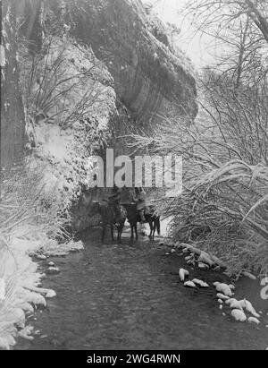 Wir nähern uns Winter-Apsaroke, 1908. Zwei Krähenindianer zu Pferd in einem flachen Bach, flankiert von schneebedeckten Bäumen, unter Felsklippen. Stockfoto