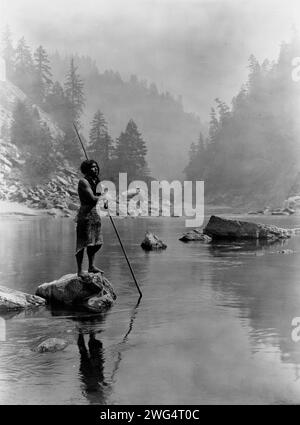 Ein rauchiger Tag im Sugar Bowl-Hupa, 1923. Hupa Mann mit Speer, stehend auf Felsen in der Mitte des Baches, im Hintergrund, Nebel verdeckt teilweise Bäume an Berghängen. Stockfoto