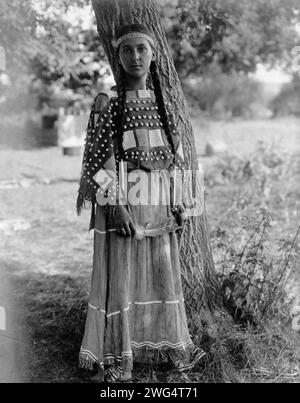 Sioux Maiden, 1908. Porträt in voller Länge, stehend am Baum, Perlenbesatz, Kleid aus perlenbesetztem Leder. Stockfoto