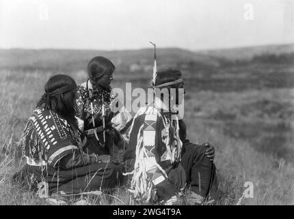 Mit Blick auf das Camp-Piegan, 1910. Ein Mann und zwei Frauen sitzen auf einem Hügel, nach rechts gerichtet. Stockfoto