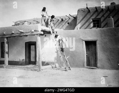 In San Ildefonso, 1927. Drei Tewa-Indianer, zwei auf dem Dach des lehmhauses, einer auf der Leiter, San Idlefonso, New Mexico. Stockfoto