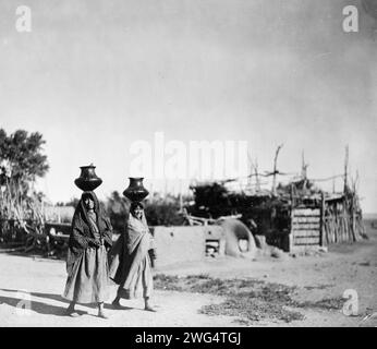 Im Dorf Santa Clara, 1905. Zwei Pueblo-Frauen, die Krüge auf dem Kopf tragen; Bäume, Zugzaun, Stockwerk entlang des unbefestigten Weges im Hintergrund. Stockfoto