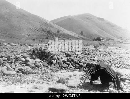 Nez PERC&#xe9; Sweat-Lodge, c1910. Landschaft, verzurrter Pol mit Decke, große glatte Steine im trockenen Flussbett, Haus, Zaun und Hügel im Hintergrund. Stockfoto