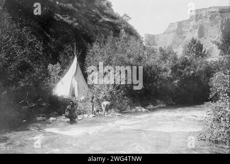 Das Flusslager, 1905. Krähenmann mit zwei Kindern und Frau (?) Am Ufer des Flusses; Tipi, umgeben von Bäumen und Bergen. Stockfoto