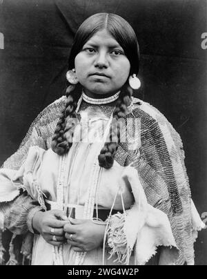 Cayuse Woman, halblanges Porträt, stehend, nach vorne gerichtet, Zöpfe, Ohrringe mit Muschelscheibe, Choker mit Muschelperlen und Muschelperlen um den Hals, Kleid aus perlenbesetztem Buckskin mit perlenbesetztem Gürtel, C1910. Stockfoto