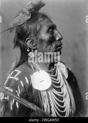 Arzneimittel Crow-Apsaroke, 1908. Medizin Crow, Crow Indian, Montana, Kopf-und-Schultern-Porträt, nach rechts gerichtet, Kopfschmuck aus hawk-Fell, zwei Ohrringe im rechten Ohr, Scheibenschmuck, Muschelperlen, Kopfschwartenhemd mit Perlen. Stockfoto