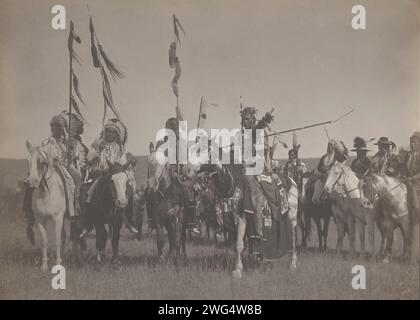 Sieg, 1908. Das Foto zeigt mehrere Krähenindianer, darunter viele Coups, Wet, Bull Don't Fall Down und Spotted Jack Rabbit, die traditionelle Kleidung und Kopfbedeckungen tragen, während sie auf dem Pferd Stab tragen. Stockfoto