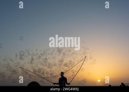 Ein Mann in der Silhouette macht viele Seifenblasen mit einem Seifenblasenstab, sie schweben am orangefarbenen Himmel bei Sonnenuntergang Stockfoto