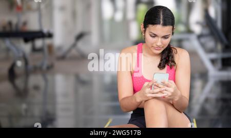Junge Frau in einem pinkfarbenen Sporttop, die ein Smartphone benutzt, während sie in einem Fitnessstudio sitzt Stockfoto