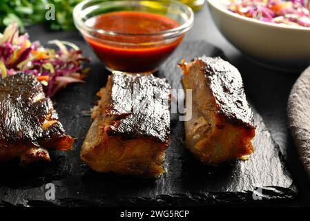 In Scheiben geschnittene scharfe gegrillte Spareribs, serviert mit Sauce auf schwarzem Schieferteller auf dunklem Hintergrund. Nahansicht Stockfoto