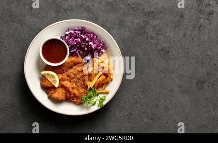 Schnitzel im Wiener Stil serviert mit Kartoffelfritten, Rotkohl-Salat und Soße auf weißem Teller über dunklem Steinhintergrund mit Kopierraum. Draufsicht, flach Stockfoto