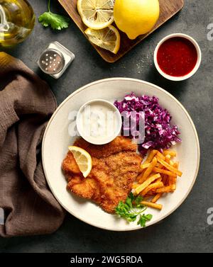 Schweineschnitzel serviert mit Kartoffelfritten, Rotkohl-Salat und Soße auf weißem Teller auf dunklem Steinhintergrund. Draufsicht, flach Stockfoto