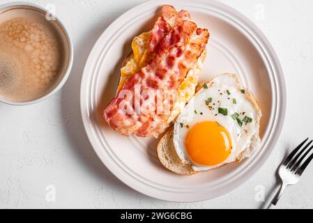 Frühstückssandwiches mit Spiegelei, Speck, Rührei und Kaffee auf weißem Tisch, Blick von oben. Spiegeleier mit Speck auf Toast und Cappuccino am Morgen Stockfoto