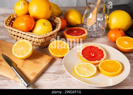 Teller mit Scheiben verschiedener Zitrusfrüchte zur Dekoration der Säfte, neben einem Schneidebrett und Grapefruits, Orangen, Zitronen und Mandarinen. Stockfoto