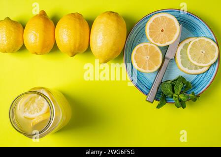 Zitronenscheiben auf einem blauen Teller mit Minze neben einer Reihe ganzer Zitronen, mit einem offenen Glas mit Zitronensaft und Scheiben darin. Stockfoto