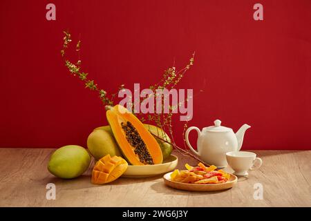 Viele Obstsorten mit etwas getrockneten Süßigkeiten auf einer Schüssel und ein Teeset auf Holztisch mit rotem Hintergrund. Vorderansicht Stockfoto