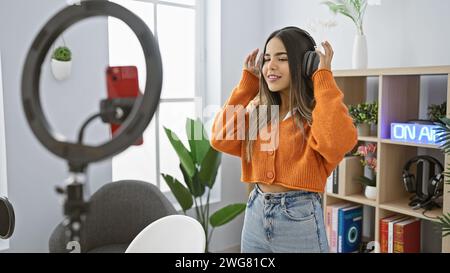 Hispanische Frau, die Musik in einem modernen Radio-Studio-Interieur genießt, mit Kopfhörern und einer Aufnahme-Ringleuchte. Stockfoto