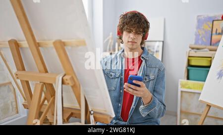 Ein junger Mann mit lockigem Haar, der eine Brille und eine Jeansjacke trägt und sein Smartphone in einem kreativen Kunststudio überprüft Stockfoto