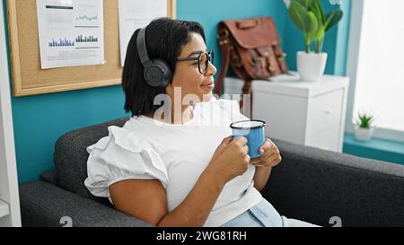 Hispanische Frau, die sich mit Kaffee und Kopfhörern in einer modernen Büroumgebung entspannt. Stockfoto