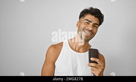 Hübscher junger hispanischer Mann, der eine Haarbürste hält, lächelt im Tanktop vor der weißen Wand Stockfoto