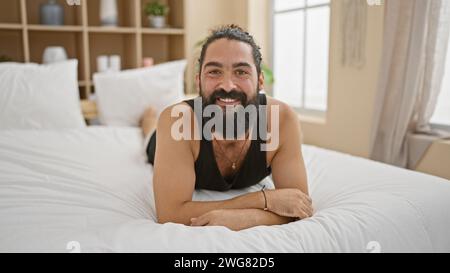 Ein fröhlicher bärtiger hispanischer Mann, der sich in einem modernen Schlafzimmer entspannt und eine ungezwungene und einladende Atmosphäre ausstrahlt. Stockfoto