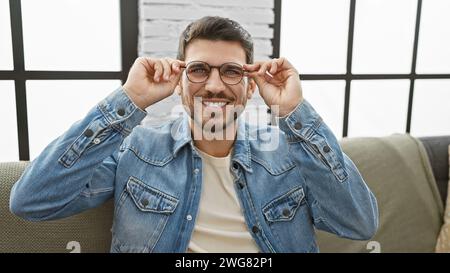 Ein hübscher junger hispanischer Mann mit Bart passt seine Brille in einem modernen Wohnzimmer an und strahlt Charisma aus. Stockfoto