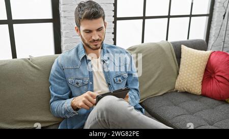 Hübscher junger hispanischer Mann mit Bart, der Denim auf der Couch trägt und drinnen Tablette benutzt. Stockfoto