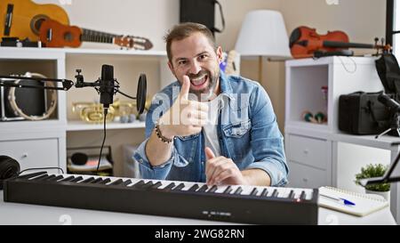 Gutaussehender reifer hispanischer Mann, der in einem Musikstudio mit Gitarren und Keyboard einen Daumen nach oben gibt. Stockfoto