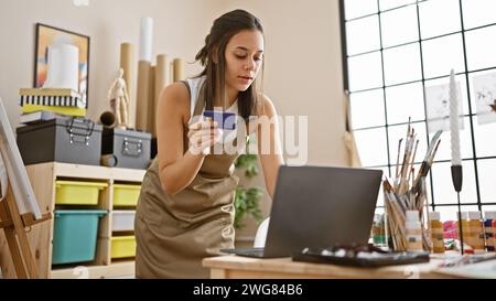 Atemberaubende junge hispanische Künstlerin, die ein Meisterwerk online im Kunststudio mit Laptop und Kreditkarte malte Stockfoto