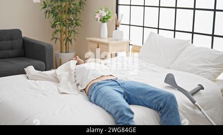 Verwungener junger Mann, der in einem modernen Schlafzimmer mit Krücken in der Nähe ruht und die Heilung zu Hause darstellt. Stockfoto