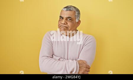 Ein nachdenklicher Mann mittleren Alters mit grauen Haaren posiert vor gelbem Hintergrund, seine Arme überkreuzt. Stockfoto