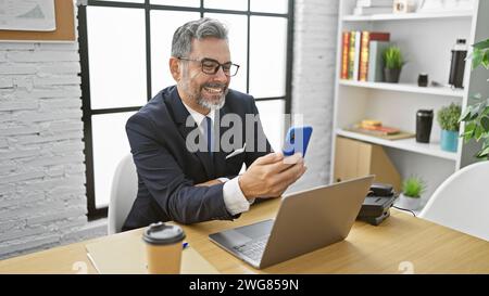 Charmanter junger hispanischer Mann, grauhaariger Angestellter, lächelnde Videoanrufe, müheloses Multitasking im Geschäft über Laptop und Smartphone aus Stockfoto