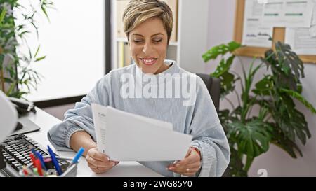 Eine lächelnde hispanische Frau mit kurzen blonden Haaren, die in einem modernen Büro ein Dokument liest. Stockfoto