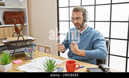 Bärtiger Mann im Büro mit Schlagstöcken, Kopfhörern, freudig, an einem modernen Arbeitsplatz mit Tageslicht Stockfoto