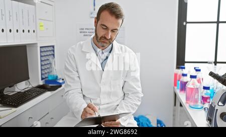 Ein hispanischer Mann mittleren Alters mit Bart, gekleidet in einem Labormantel, konzentriert sich darauf, Notizen in einem klinischen Labor zu schreiben. Stockfoto